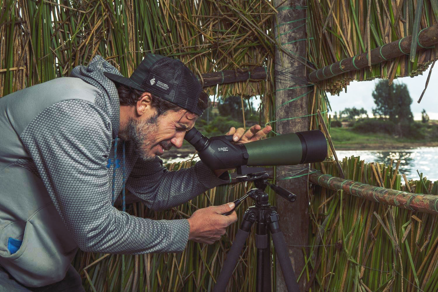 Man birdwatching through a spotting scope at Ensifera Camp in the Sacred Valley of the Incas, inside a reed observation hut overlooking the water – nature tourism and wildlife photography in Peru.