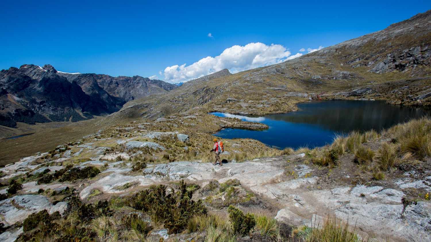 Excursionista en el Santa Cruz Trek, en la Cordillera Blanca del Perú, caminando por un terreno rocoso de altura junto a una laguna alpina azul profundo, rodeada de montañas bajo un cielo despejado.