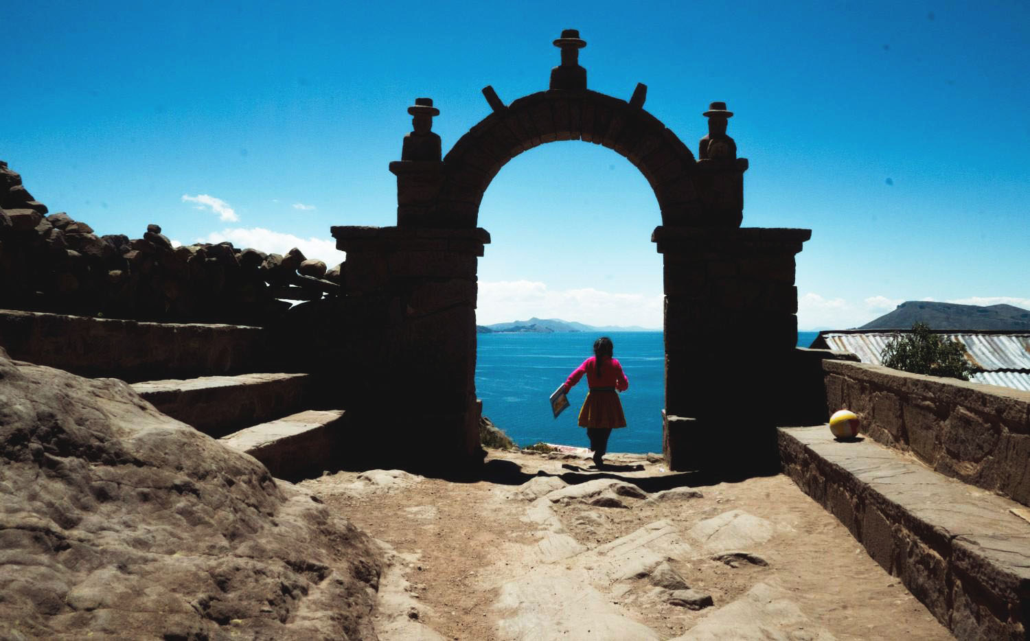 Woman walking through a traditional stone archway on Taquile Island, with Lake Titicaca altitude and its islands in the background – cultural and experiential tourism in the Peruvian highlands.