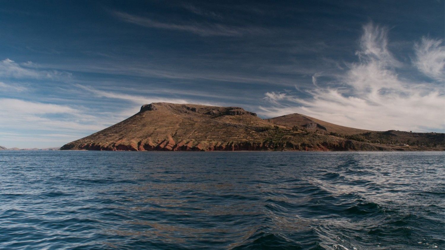 View from the lake of Amantani Island on Lake Titicaca, with its agricultural terraces and hills under an Andean sky – cultural tourism and nature in the Peruvian highlands.